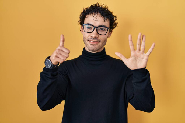 Hispanic man standing over yellow background showing and pointing up with fingers number six while smiling confident and happy.  - Foto, afbeelding