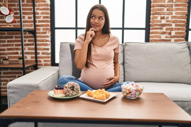 Νεαρή έγκυος γυναίκα τρώει γλυκά στο σπίτι σοβαρό πρόσωπο σκέφτεται την ερώτηση με το χέρι στο πηγούνι, στοχαστική για σύγχυση ιδέα  - Φωτογραφία, εικόνα
