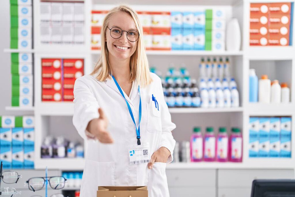 Joven mujer rubia farmacéutica sonriendo con confianza estrechan la mano en la farmacia - Foto, imagen