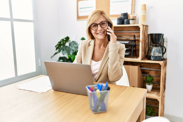 Středního věku blondýny žena usmívá sebevědomě mluvit na smartphone v kanceláři - Fotografie, Obrázek