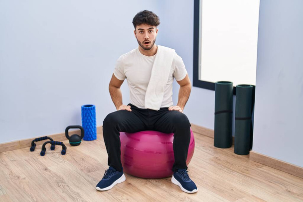 Hispanic man with beard sitting on pilate balls at yoga room afraid and shocked with surprise expression, fear and excited face.  - Zdjęcie, obraz