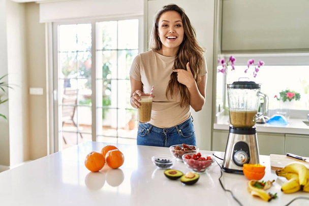 Gyönyörű fiatal barna nő iszik üveg smoothie a konyhában ujjal mutogatva egy önmosolygó boldog és büszke  - Fotó, kép