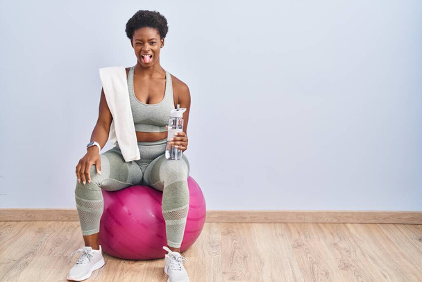 Afro-Amerikaanse vrouw draagt sportkleding zittend op pilates bal steekt tong uit gelukkig met grappige uitdrukking. emotie concept.  - Foto, afbeelding