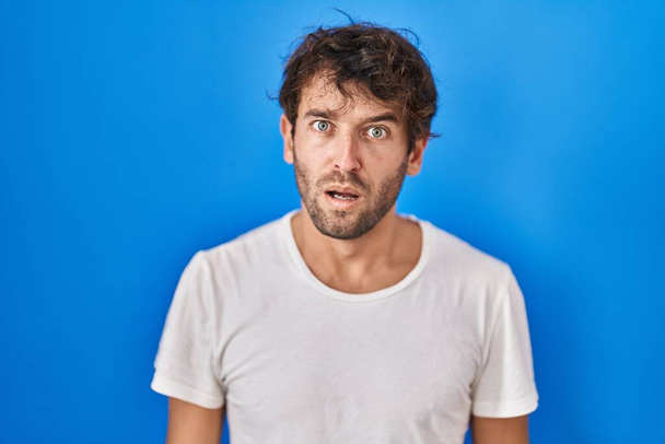 Hispanic young man standing over blue background in shock face, looking skeptical and sarcastic, surprised with open mouth  - Photo, Image