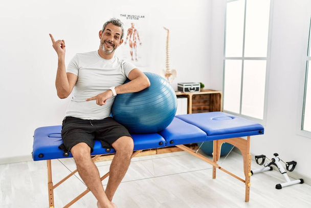 Hombre hispano de mediana edad en la clínica de recuperación del dolor sosteniendo una pelota de pilates con una gran sonrisa en la cara, señalando con el dedo de la mano hacia el lado mirando a la cámara.  - Foto, Imagen