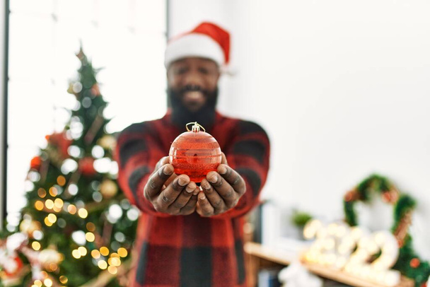 Giovane africano americano uomo sorridente fiducioso tenendo palla in piedi dal cappello di Natale a casa - Foto, immagini