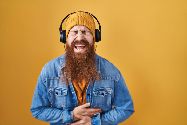 Kaukasischer Mann mit langem Bart, der über Kopfhörer Musik hört, lächelt und laut lacht, weil er einen lustigen verrückten Witz mit den Händen am Körper macht.  - Foto, Bild