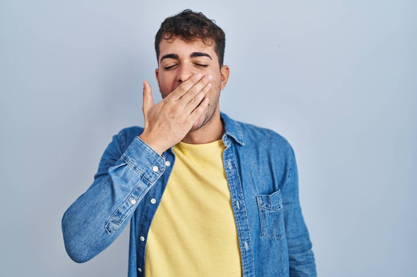 Jeune homme hispanique debout sur fond bleu ennuyé bâillement fatigué couvrant la bouche avec la main. agitation et somnolence.  - Photo, image