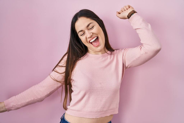 Giovane donna bruna in piedi su sfondo rosa che balla felice e allegra, sorridendo commovente casuale e fiducioso ascoltando musica  - Foto, immagini