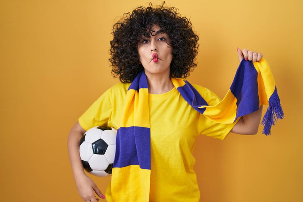jonge brunette vrouw met krullend haar voetbal hooligan holding bal het maken van vis gezicht met mond en squinting ogen, gek en komisch.  - Foto, afbeelding