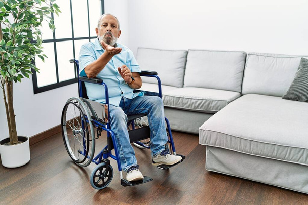 Pohledný starší muž sedící na vozíčku v obývacím pokoji a dívající se do kamery, který posílá polibek s rukou na vzduchu, je rozkošný a sexy. výraz lásky.  - Fotografie, Obrázek