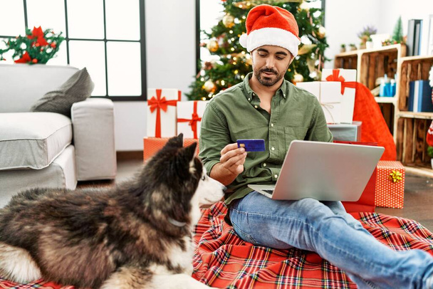 Młody Hiszpan za pomocą laptopa i karty kredytowej siedzi na podłodze z psem przez choinkę w domu - Zdjęcie, obraz