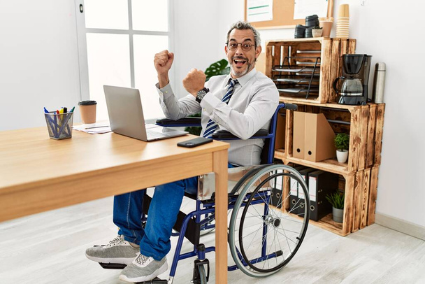 Μεσήλικας Ισπανός που εργάζεται στο γραφείο και κάθεται σε αναπηρική καρέκλα γιορτάζοντας έκπληκτος και έκπληκτος για την επιτυχία με τα χέρια σηκωμένα και ανοιχτά μάτια. έννοια νικητή.  - Φωτογραφία, εικόνα