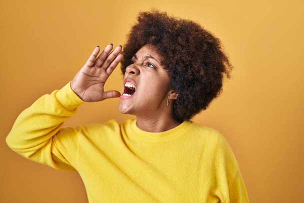 Jonge Afro-Amerikaanse vrouw die over een gele achtergrond staat schreeuwend en schreeuwend met hand op mond. communicatieconcept.  - Foto, afbeelding