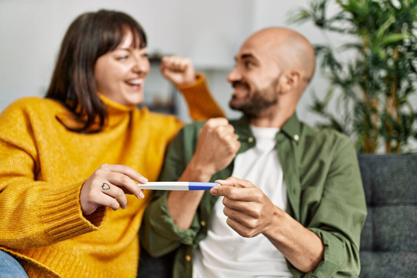 Young hispanic couple smiling happy holding pregnancy test sitting on the sofa at hom - Foto, Imagen
