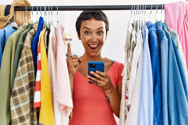 Молода іспаномовна жінка з коротким волоссям шукає одяг на стійці для одягу, використовуючи смартфон, здивований ідеєю або питанням, спрямованим пальцем з щасливим обличчям, номер один
  - Фото, зображення