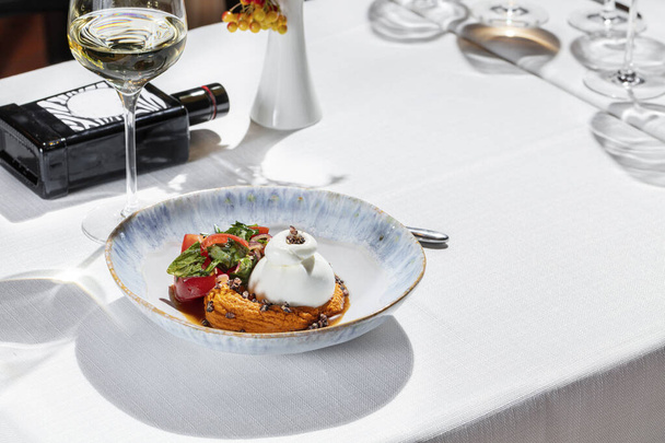 Buffalo mozzarella on lentil puree with finely chopped olives, next to a salad of tomatoes and herbs. Food is in a light ceramic plate, next to it is a glass of white wine and a bottle of olive oil. - Photo, Image