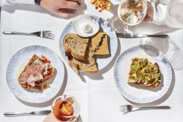En una mesa con un mantel ligero con bruschettas con jamón y alcachofas, foie gras paté y copas de rosa y vino blanco, un hombre y una mujer están sentados. - Foto, imagen