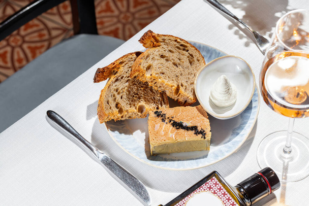 Paté de Foie gras con aceite de oliva y aceitunas pequeñas. pan de trigo sarraceno y salsa de crema. Comida en un plato de cerámica ligera sobre un mantel ligero. Cerca hay una copa de vino de rosas, un jarrón y cubiertos.. - Foto, imagen