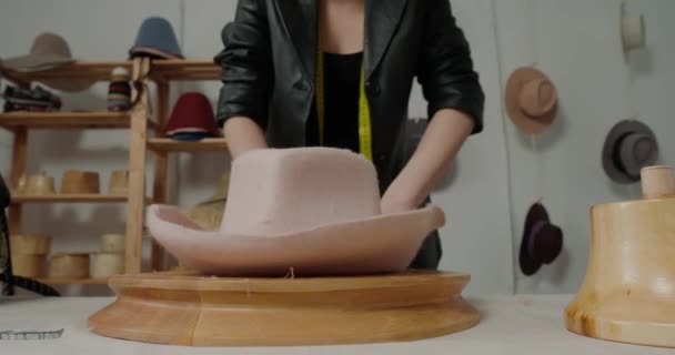 Woman in leather jacket makes shape of handmade felt hat on wooden mold at table. Professional milliner creates stylish headwear in workshop - Кадры, видео