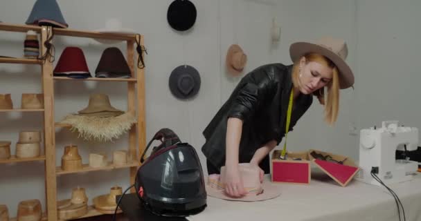 Stylish woman in leather jacket decorates felt hat with beads and ribbon standing at table with equipment. Process of making headwear in workshop - Кадры, видео