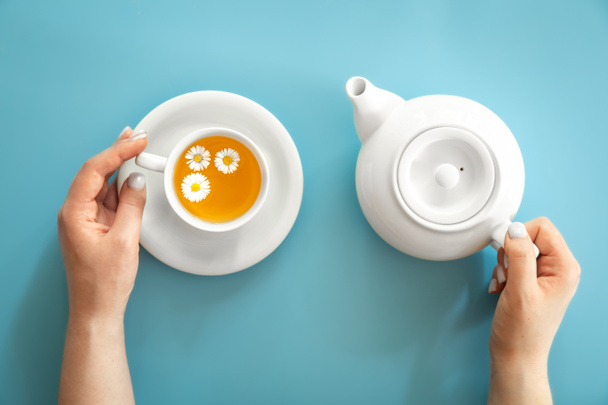 A cup of chamomile tea and a teapot in female hands on a blue background, flat lay, minimalism. - Fotoğraf, Görsel