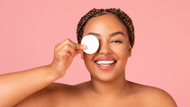 Facial Skincare. Pretty Overweight African American Lady Covering Eye With Cotton Pad Posing Smiling To Camera Over Pink Studio Background. Beauty And Skin Care Cosmetics Concept. Panorama - Foto, immagini