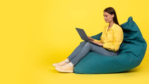 Side View Of Female Using Laptop Working Online Browsing Internet Sitting In Beanbag Chair Posing In Studio Over Yellow Background. Web Technology Concept. Panorama With Copy Space - Fotoğraf, Görsel