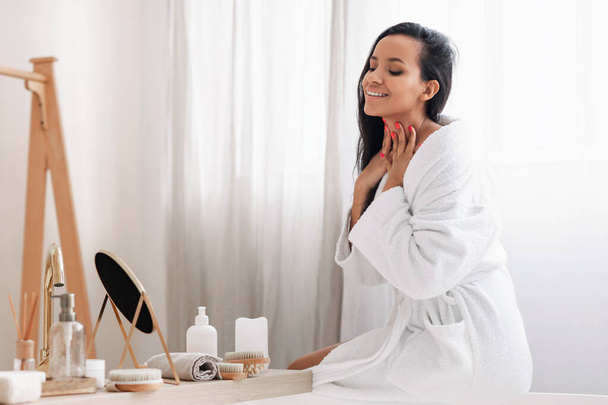 Pretty Mixed Woman Touching Neck Caring For Skin Looking At Mirror Enjoying Beauty Routine Sitting In Modern Bathroom Indoors. Skincare Cosmetic Products Concept - Φωτογραφία, εικόνα