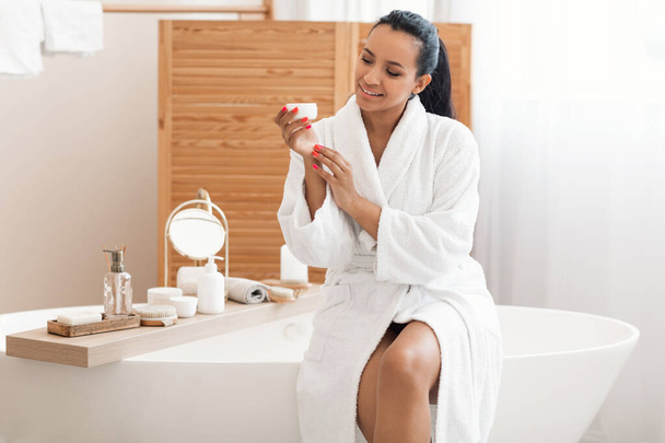 Skincare Cosmetics. Mixed Female Holding Moisturizer Jar Moisturizing Hands Skin Sitting In Modern Bathroom, Wearing White Bathrobe. Beauty Care Routine Concept - Fotó, kép