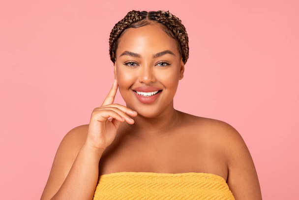 Portrait Of Attractive Obese Black Woman Touching Face Posing Smiling To Camera Over Pink Studio Background, Standing Wrapped In Towel. Beauty, Wellness And Spa Concept - Фото, изображение