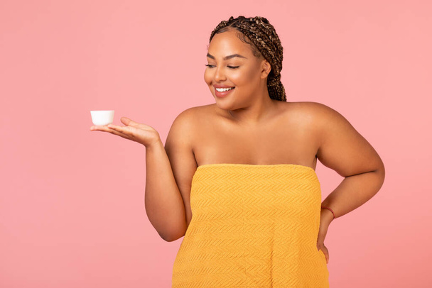 Pretty Obese Black Woman Holding Moisturizer Cream Jar Posing Wrapped In Towel Caring For Skin Standing Over Pink Studio Background. Facial Skincare Cosmetics, Beauty Routine Concept - 写真・画像