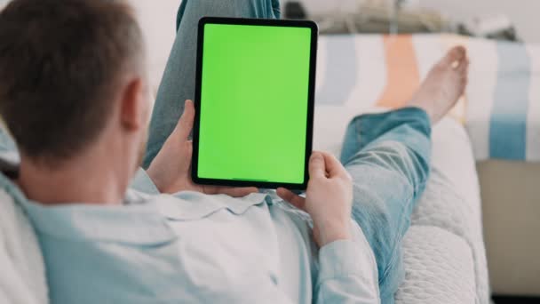 man nodding head lying on a couch wearing casual shirt using digital tablet with green screen for video call leisure time at home - Filmati, video