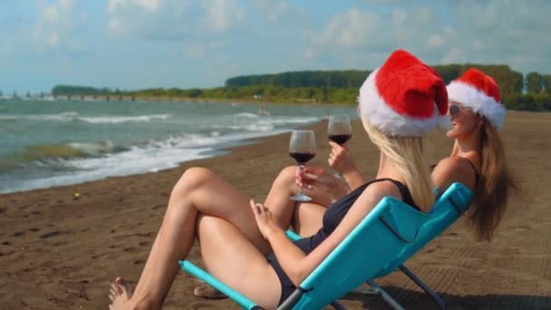 Happy girls in bikinis sit on chairs celebrating Christmas on beach, drinking wine in santa's red hats - young smiling woman - Christmas beach party concept. Hit glasses. coming new year - Footage, Video