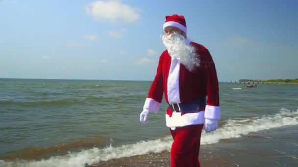 Санта-Клаус в костюме прогуливается вдоль берега моря или океана. Экзотическая встреча Рождества на острове на пляже с песком в жаркую погоду или экзотический климат. Волны, море. Время отдыхать в отпуске. - Кадры, видео