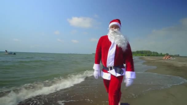 Santa Claus in suit walks and jumps along sea or ocean shore. Exotic Christmas encounter on island on beach with sand in hot weather or exotic climate. Waves, sea. Time to relax on vacation. - Кадры, видео