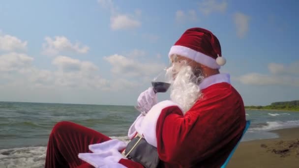 Kostümlü Noel Baba deniz ya da okyanus kıyısındaki sahilde camdan kırmızı şarap içer. Noel 'i sıcak iklimde egzotik bir ülkede kutluyor. Yılbaşından önce dinlen ve eğlen. Rahatlama ve mutluluk. - Video, Çekim