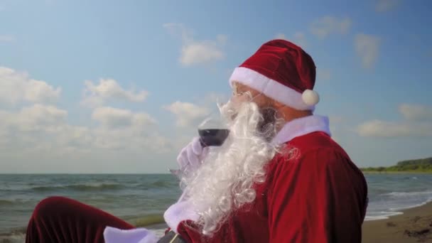 Санта-Клаус в костюме пьет красное вино из бокала на пляже возле моря или океана. Он празднует Рождество в экзотической стране в жарком климате. Отдых и веселье перед Новым годом. Расслабление и счастье. - Кадры, видео