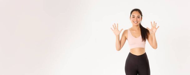 Sport, Wohlbefinden und aktiver Lebensstil. Lächelnd starke und schlanke asiatische Fitnesslehrerin, die Lektion erteilt, erklärt zehn Schritte für perfekten Körper und produktives Workout, weißer Hintergrund. - Foto, Bild