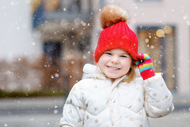 Funny portrait of little preschool girl in winter clothes. Happy positive child with red hat and colorful gloves outdoors. Winter day - Photo, Image