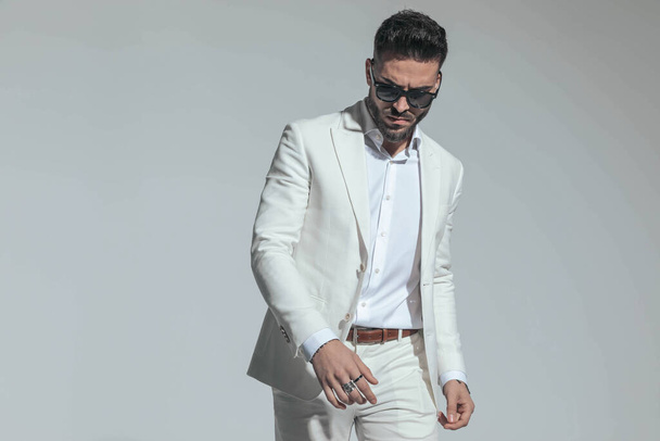 portrait of sensual smart casual man in white suit looking down and posing in a cool manner on grey background - Fotó, kép