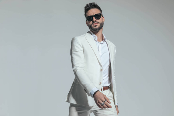 portrait of unshaved cool man wearing shite stylish suit holding arms in fashion pose, being confident and posing in front of grey background - Photo, image
