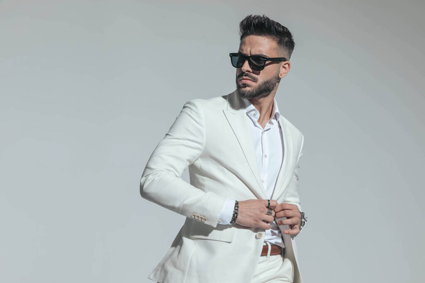 portrait of sexy young man in white suit with open collar shirt buttoning jacket and looking to side while posing in a cool manner on grey background - Fotó, kép