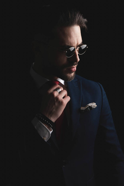 cut out picture of handsome fashion man with retro sunglasses fixing tie and looking to side while posing in a mysterious way on black background  - Foto, afbeelding