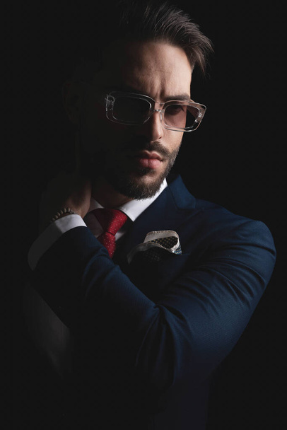 cut out picture of seductive businessman in elegant suit with sunglasses touching neck and looking to side on black background in studio - Foto, afbeelding