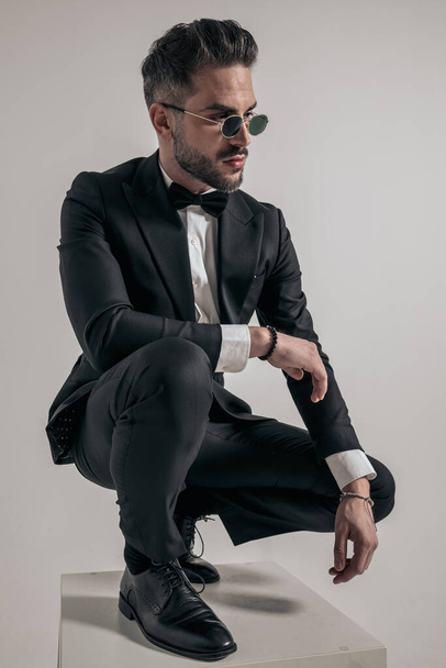 sexy elegant man in tuxedo with bowtie crouching and looking to side while holding elbow on knee on grey background - Φωτογραφία, εικόνα