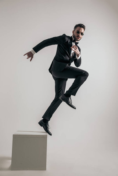 side view of bearded man in tuxedo jumping to new beginnings and new opportunities on grey background - Foto, afbeelding