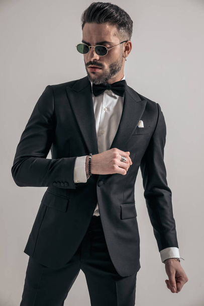portrait of sexy bearded guy in black tuxedo holding arms in fashion pose and looking away in studio - Photo, Image