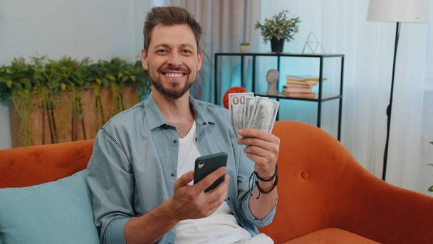 Planning family budget. Smiling happy man counting money dollar cash, use smartphone calculate domestic bills at home. Joyful young guy satisfied of income saves money for planned vacation, gifts - Foto, Bild