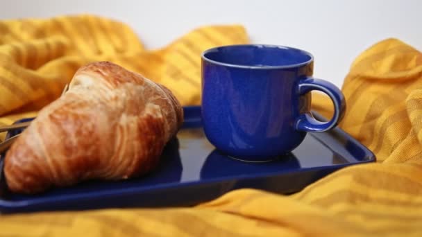 Blue steaming mug and plate with hot french baked croissant stand on a orange cloth - prepared for a regular customer in coffee house and bakery. Breakfast or coffee break in cafe. No people on video - Кадры, видео